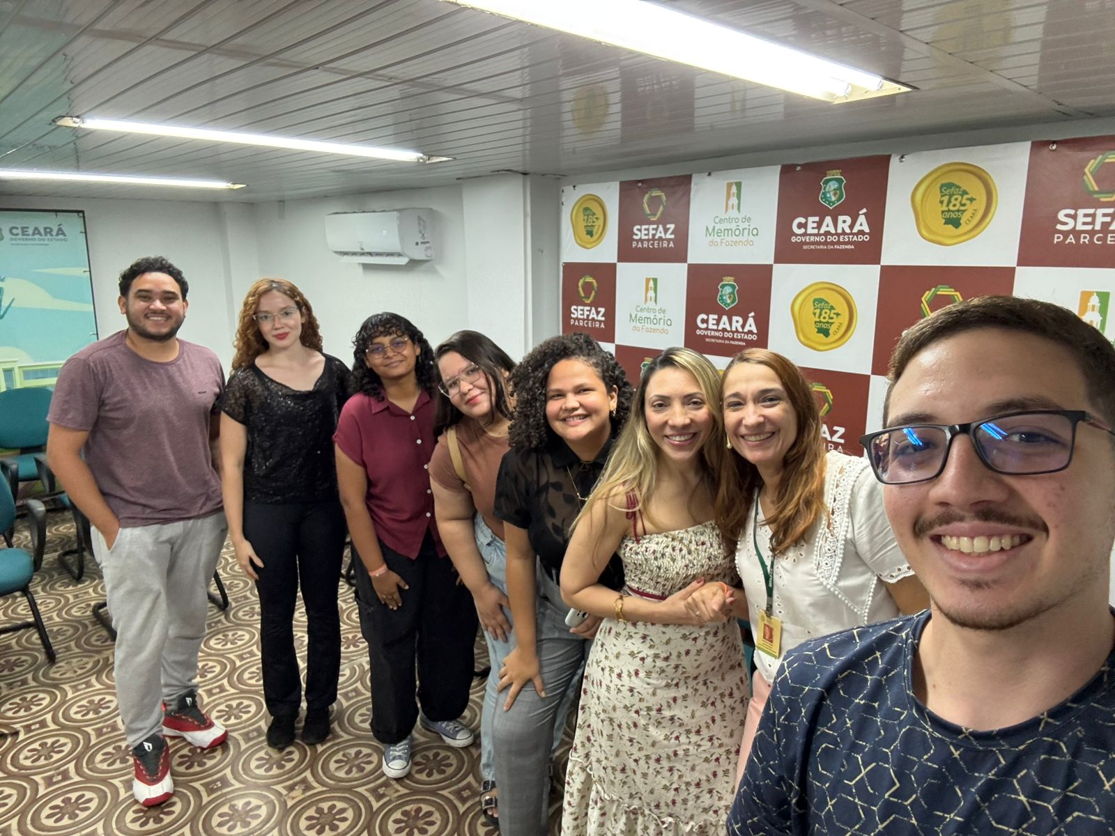 Na imagem, oito pessoas posam para uma selfie em uma sala, todas sorrindo em direção à câmera. O homem à direita segura o celular, enquanto os demais integrantes do grupo estão alinhados atrás dele. Ao fundo, vê-se um painel com as logomarcas do Governo do Estado do Ceará, da Sefaz e do Centro de Memória da Fazenda, além de um ar-condicionado fixado na parede. O chão possui um padrão geométrico em tons claros. Todos aparentam estar descontraídos e felizes, formando um ambiente acolhedor e amistoso