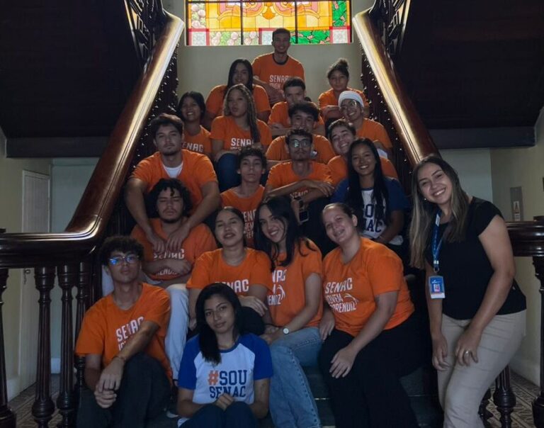 Foto de grupo com a professora Eleni Sousa e sua turma do curso de Auxiliar Administrativo da Instituição Senac em uma escadaria, em frente a um vitral. Todos estão sorrindo, e a maioria usa camisetas laranjas. A foto é datada de 18 de julho.