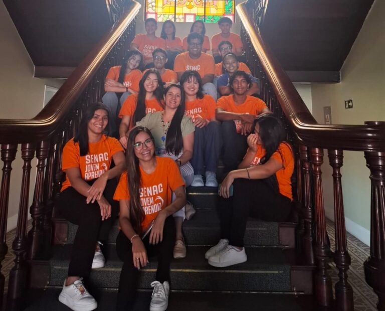 A imagem mostra um grupo de 15 a 20 estudantes e uma professora, todos vestindo camisetas laranjas do Senac, sentados em uma grande escadaria de madeira. No topo da escada, há um grande vitral colorido. A professora Luana Siqueira está sentada no centro da foto, com um sorriso. O grupo está olhando para a câmera com expressões felizes