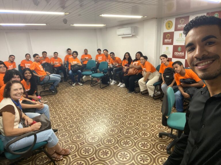 Selfie de um grupo de cerca de 20 pessoas, incluindo a professora Rosângela Xavier, em uma sala de aula no dia 25 de julho. A maioria dos participantes, que são jovens, está vestindo camisetas laranja. O grupo está sentado em cadeiras em um semicírculo. Ao fundo, banners com os logos da Sefaz Ceará e do Centro de Memória estão visíveis. A foto documenta a visita ao Centro de Memória da Fazenda.