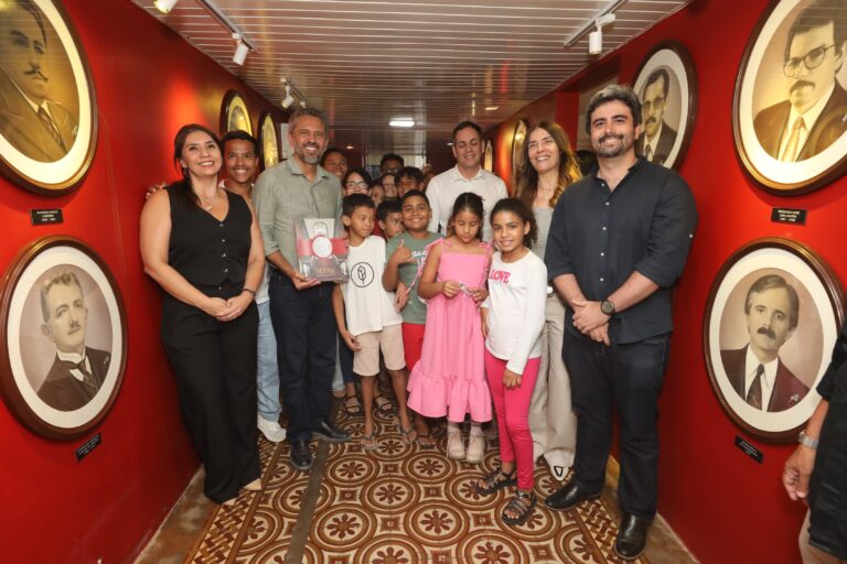 Fotografia tirada em 17 de julho. O governador Elmano de Freitas posa em um corredor com paredes vermelhas no Centro de Memória da Fazenda. Ele está ladeado por adultos e crianças. A parede está decorada com retratos redondos em preto e branco.