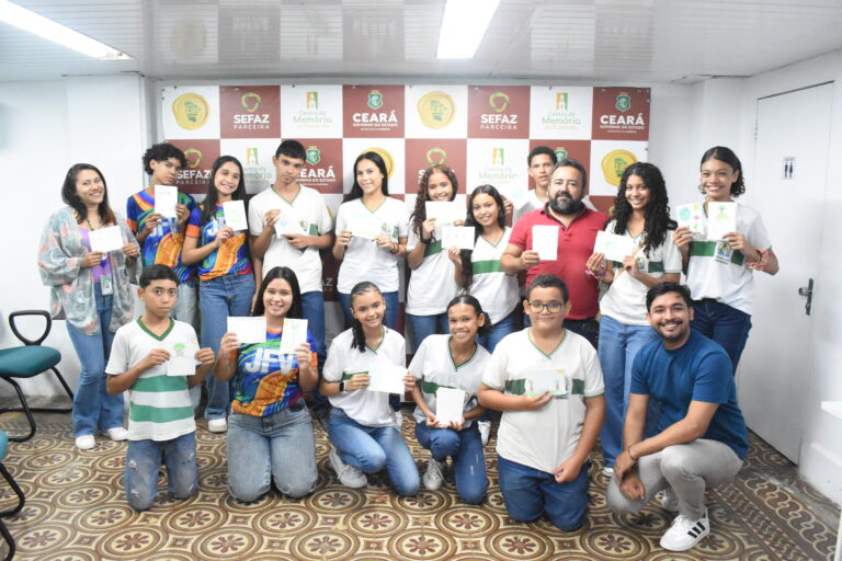 Um grupo de estudantes posa sorridente em frente a um painel com os logotipos do Governo do Ceará, da Sefaz e do Centro de Memória da Fazenda. Eles exibem trabalhos em papel que parecem desenhos ou produções escolares. Dois educadores acompanham o grupo, posicionados nas laterais.