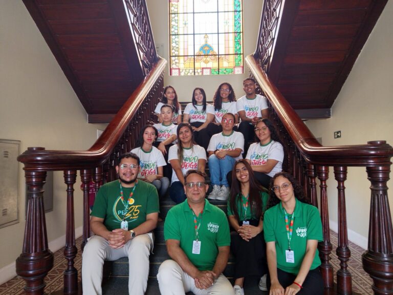 Grupo de 12 pessoas, incluindo nove alunos e três monitores (Jean Dumont, Natielly Oliveira e Gabriele Oliveira), sentados e em pé em uma escadaria de madeira. O grupo usa camisetas do projeto "Virando o Jogo" e posa para uma foto. Um vitral colorido é visível ao fundo.