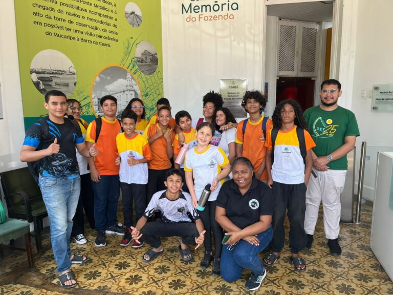 Um grupo de 17 pessoas, sendo 15 alunos e a professora Flávia Sousa, posam para uma foto em pé, na entrada de um prédio. Ao fundo, um painel com a logo "Centro de Memória da Fazenda" é visível.