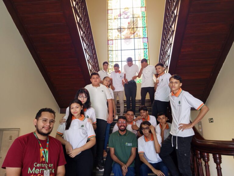 Um grupo de 16 pessoas, incluindo alunos em uniforme escolar e um professor, posa para uma foto em uma escadaria de madeira. O professor, que usa uma camisa polo verde, está na frente do grupo.