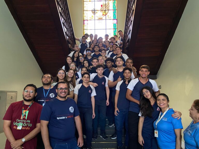Grande grupo de alunos e professores do Colégio Ateneu, todos com uniformes azuis, posam para uma foto em uma escadaria de madeira, com um vitral colorido ao fundo.