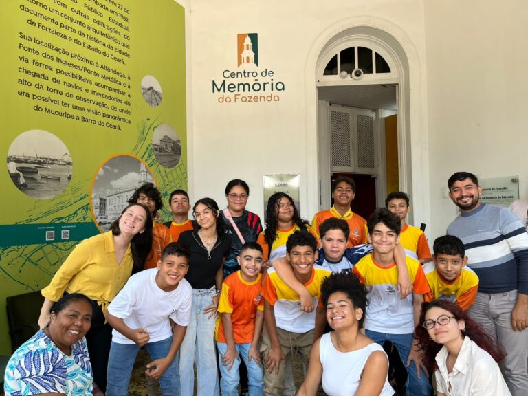 Um grupo de 17 pessoas, sendo 15 alunos e a professora Flávia Sousa, posam para uma foto em pé, na entrada de um prédio. Ao fundo, um painel com a logo "Centro de Memória da Fazenda" é visível.