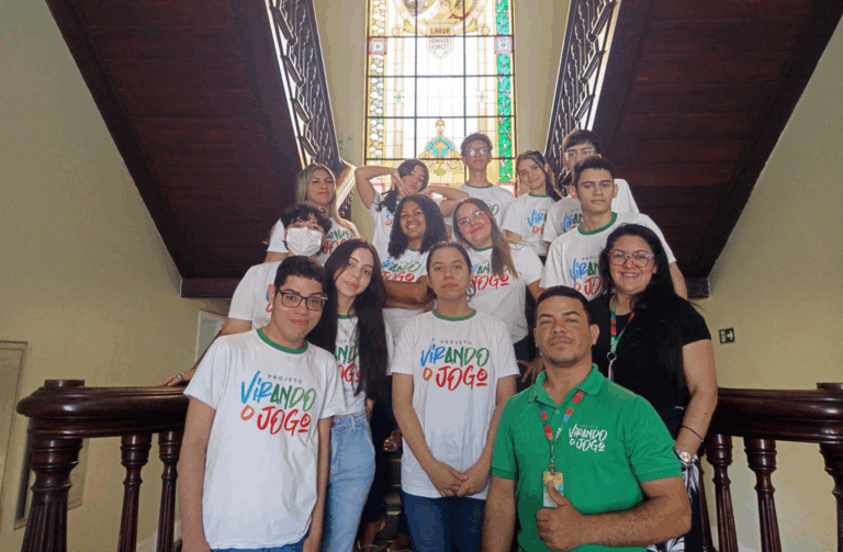 Um grupo de jovens e adultos posa em uma escadaria de madeira, em frente a um vitral colorido com a inscrição "Labor Omnia Vincit". Eles vestem camisetas brancas do *Projeto Virando o Jogo*, enquanto dois adultos usam crachás de identificação e acompanham o grupo.