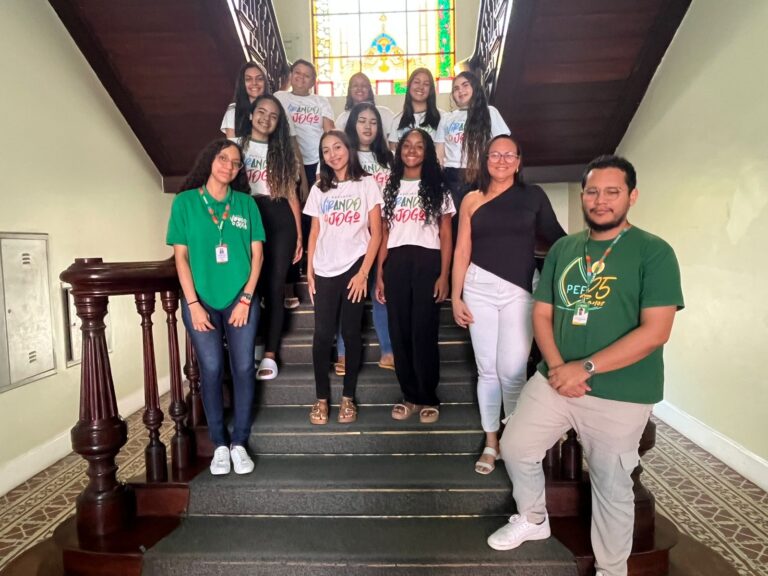 Grupo de 12 pessoas, incluindo a professora Rosi Albuquerque e alunos do projeto Virando o Jogo, posam em uma escadaria interna de prédio histórico, com vitral colorido ao fundo.
