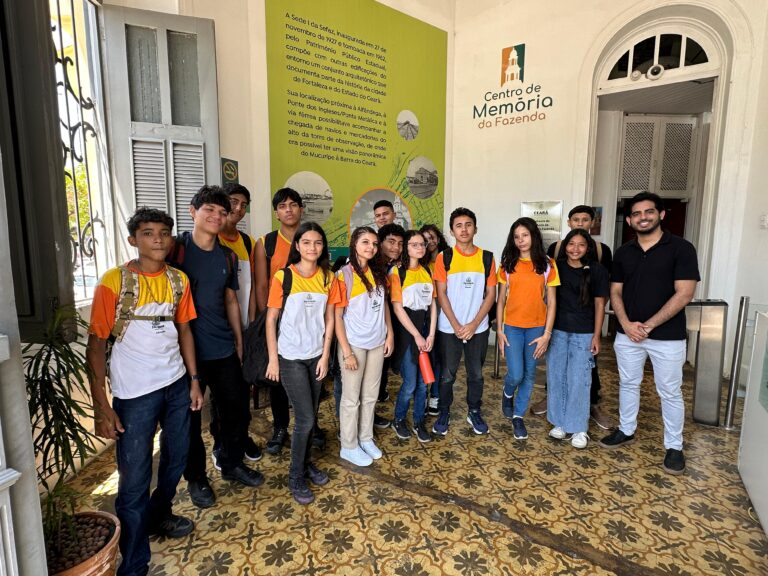 Grupo de 15 alunos da Escola Agerson Barbosa, acompanhados pelo professor Samuel Paz, posam para foto no hall de entrada do Centro de Memória da Fazenda, diante de um painel explicativo.