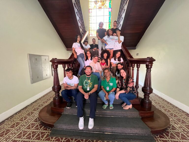 Grupo de 15 pessoas, entre alunos do projeto Virando o Jogo e a professora Nayanne Abreu, posam sorridentes em uma escadaria de madeira com vitral colorido ao fundo.