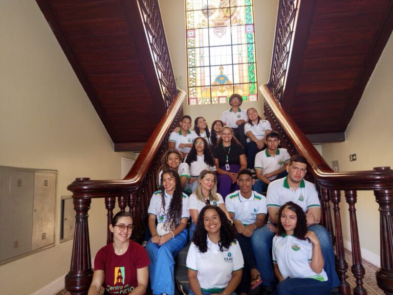 Grupo de alunos e professoras da EEMTI Rotary Club posam na escadaria interna do Centro de Memória da Fazenda, com vitral colorido ao fundo e educadores do espaço ao lado.