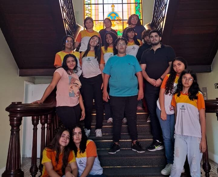 Grupo de 15 alunos da Escola Agerson Tabosa e o professor Daniel posam em uma escadaria sob um vitral durante visita ao Centro de Memória da Fazenda.
