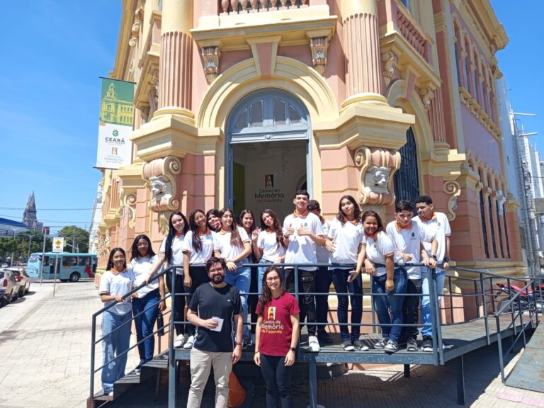 Grupo de alunos e professor da EEMTI Raul Tavares Cavalcante posam na entrada do prédio histórico do Centro de Memória da Fazenda, sob o sol da manhã.