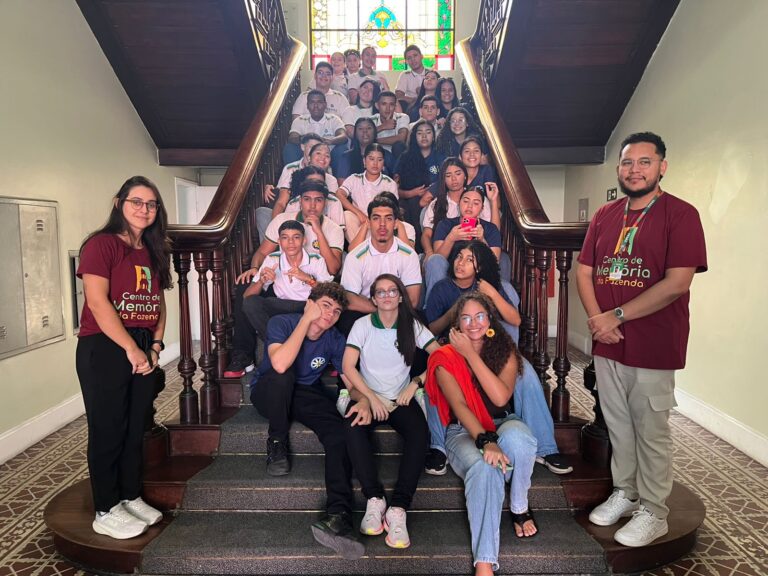 Grupo de alunos e professora da EEMTI Telina Barbosa posam sorridentes na escadaria interna do Centro de Memória da Fazenda, com vitral colorido ao fundo.