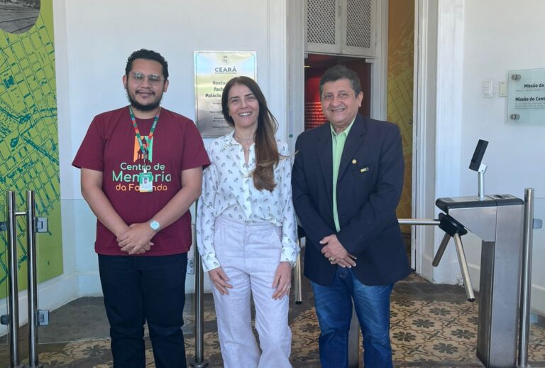 Três pessoas posam sorrindo na entrada do Centro de Memória da Fazenda durante a visita do vereador Gerôncio Coelho.