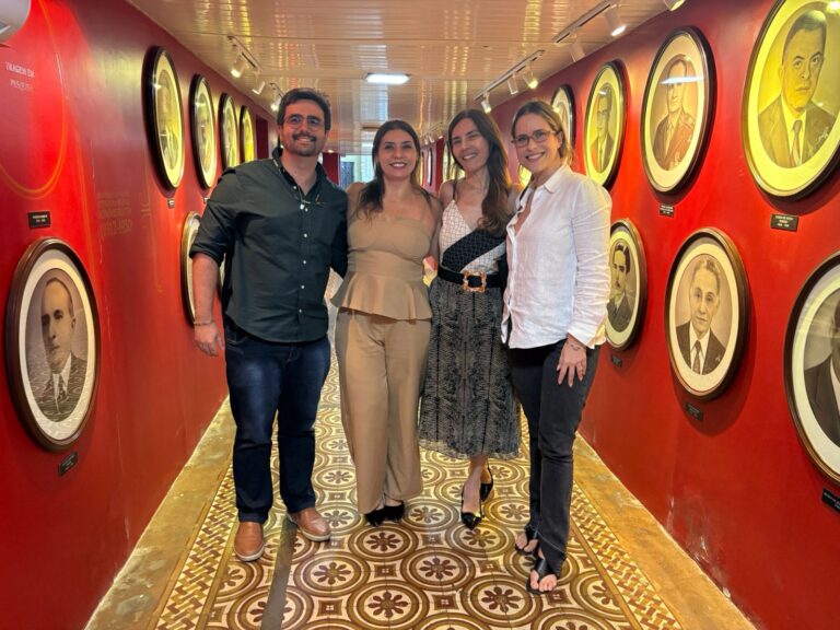 Cecília Moreira da Rocha posa ao lado de três pessoas no corredor do Centro de Memória da Fazenda, ladeado por retratos de ex-secretários da Sefaz-CE emoldurados sobre parede vermelha.