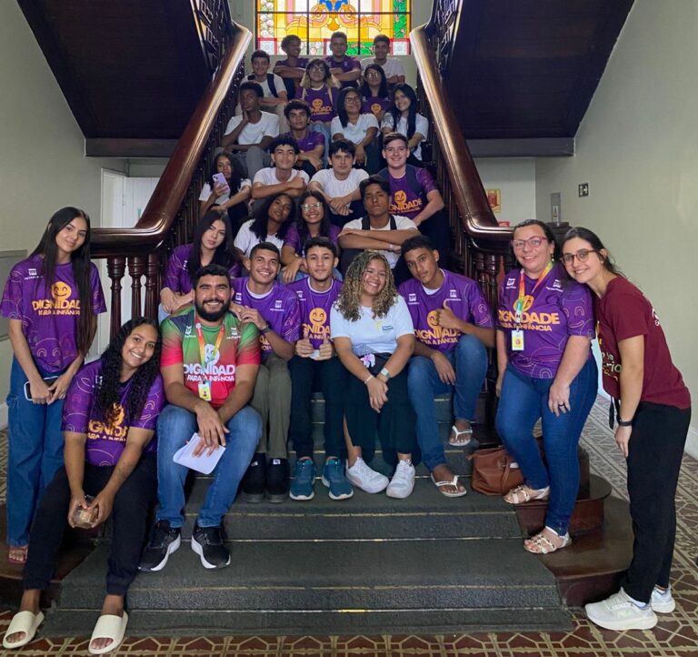 Um grande grupo de 26 alunos e o professor Aldair Alves, do projeto O Pequeno Nazareno, posando na escadaria central da SEFAZ, com muitos alunos vestindo camisetas roxas com a palavra "DIGNIDADE".
