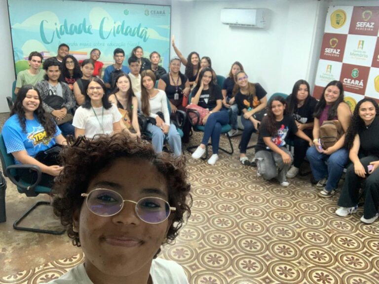 Foto de um grupo de 28 alunos do CIEE reunidos em uma sala do projeto Cidade Cidadã, acompanhados da professora Cíntia Muniz. Os estudantes estão sentados e sorrindo para a câmera enquanto uma aluna na frente faz uma selfie. Ao fundo, há um painel com a frase “Cidade Cidadã – Aprenda mais sobre educação fiscal” e, ao lado, um backdrop com logos da Sefaz, Centro de Memória e Governo do Ceará. O ambiente é iluminado e organizado com cadeiras dispostas em círculo.