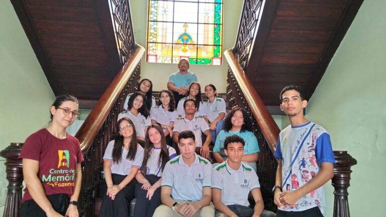 Foto de um grupo de 12 alunos da EEMTI Vicente Arruda posando em uma escadaria de madeira, com um grande vitral colorido ao fundo. Os estudantes estão distribuídos pelos degraus, sorrindo para a câmera. À esquerda está uma integrante da equipe do Centro de Memória, e à direita um aluno em pé. No topo da escadaria, um homem está sentado com as mãos apoiadas sobre os joelhos. A iluminação natural destaca o vitral e as madeiras escuras da estrutura.