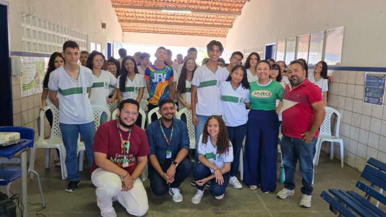 Foto de um grande grupo de estudantes da EMEF José Fernandes Vieira, em Maranguape, posando para a câmera após uma palestra de Educação Fiscal. Na frente, três membros da equipe do CM estão agachados, sorrindo, enquanto professores e mais alunos se posicionam ao fundo, todos uniformizados. O ambiente é um corredor amplo da escola, com cadeiras brancas ao redor e cartazes informativos nas paredes.