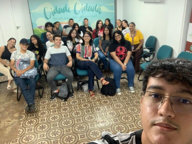 Foto de um grupo de 18 alunos do CIEE sentados em cadeiras em uma sala de aprendizagem, acompanhados da professora Cíntia Muniz. Todos estão olhando para a câmera enquanto um dos alunos tira a selfie. Ao fundo, há um painel com a frase “Cidade Cidadã – Aprenda mais sobre educação fiscal”. O grupo está sorridente e reunido de forma descontraída.