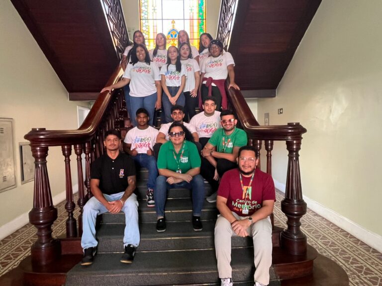 Quatorze alunos e o professor Paulo Henrique, do projeto Virando o Jogo, posando em uma escadaria de madeira, com a maioria dos alunos vestindo camisetas brancas com a logo do projeto.