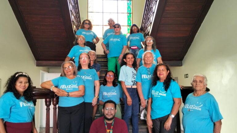 Grupo de 15 pessoas, principalmente idosas, da ABEMCE, posando em uma escadaria de madeira. A maioria veste camisetas azuis-turquesa com a frase "Feliz Idade".