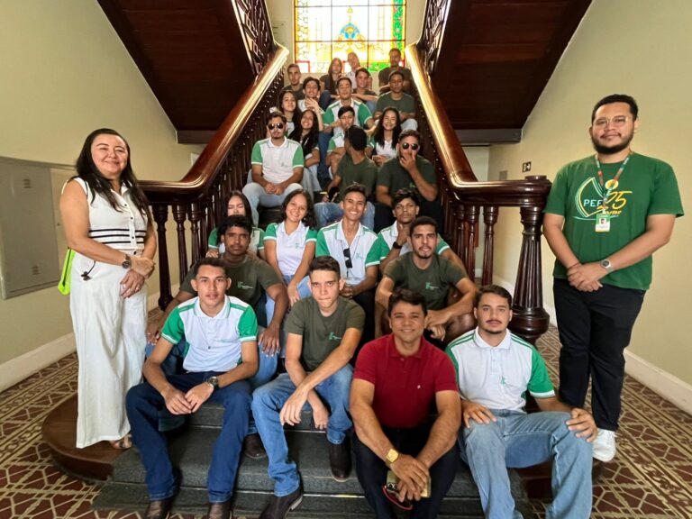 Foto em grupo de aproximadamente 28 a 30 pessoas (incluindo alunos e orientadores) reunidas em uma escadaria histórica interna, com corrimão de madeira e um vitral colorido no topo. A maioria dos alunos veste camisetas com as cores verde e branco. Duas pessoas se destacam nas laterais: uma mulher à esquerda, de vestido branco, e um homem à direita, de camiseta verde escura com o logo "PFE". A imagem registra a visita de alunos do IFTO (Instituto Federal do Tocantins).