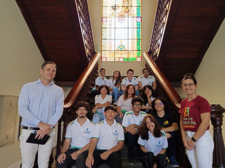 Alunos da EEMTI Anísio Teixeira e a professora Vitória Jucá, acompanhados por um funcionário da SEFAZ, posando na escadaria histórica, com um vitral colorido ao fundo.