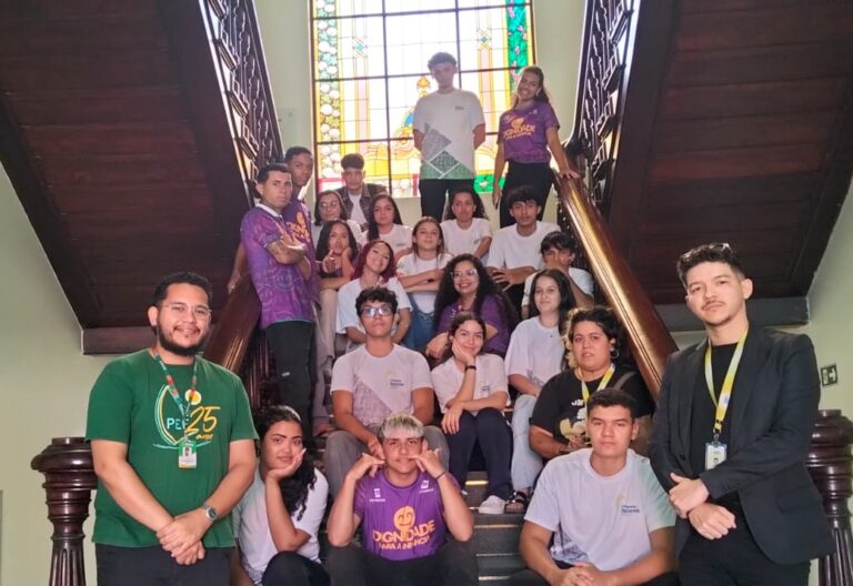 Grande grupo de 30 alunos do projeto O Pequeno Nazareno, acompanhados pelos professores Gutemberg Oliveira e Creliane Marreiro, posando em uma escadaria de madeira, com alguns alunos vestindo camisetas roxas e outros camisetas brancas.