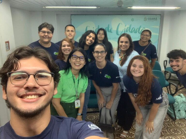 Selfie em grupo de 12 pessoas (10 alunos e 2 professoras), sorrindo em um ambiente interno. A maioria usa camisetas azuis escuras e está reunida em torno de duas mulheres vestindo blusas de cores claras (verde e azul claro). Ao fundo, há uma parede com um banner que exibe a palavra "Cidadão" e o logo do governo do Ceará. A foto foi tirada após uma atividade do NAF-Uece em 29/10 (tarde).
