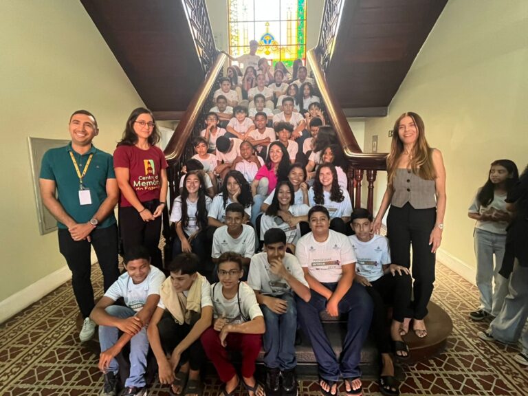 Grupo de 69 alunos e três adultos, incluindo o Professor Leonardo Alves, posando em uma escadaria interna de madeira. Os alunos, a maioria adolescentes, vestem camisetas brancas do programa, e estão sentados e em pé degrau a degrau. Os três adultos estão nas laterais. O piso térreo tem um padrão de ladrilhos coloridos. A iluminação é natural, vinda de uma janela no topo da escada, que possui um vitral.