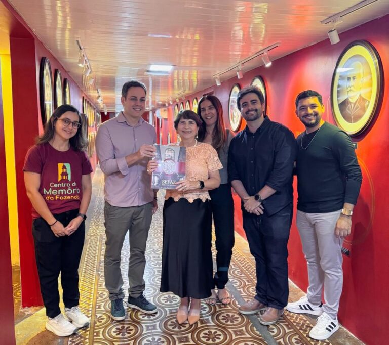 Seis pessoas posam para foto em um corredor vermelho iluminado, decorado com retratos circulares nas paredes. O grupo segura um livro no centro da imagem e sorri para a câmera.