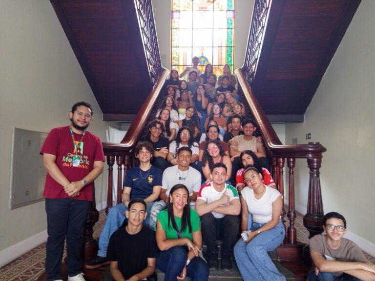 Grupo grande de adolescentes e jovens adultos posa sentado na escadaria interna de um prédio histórico, com vitral colorido ao fundo. Eles estão distribuídos pelos degraus, acompanhados por dois adultos — um homem à esquerda vestindo camisa vinho e uma mulher à frente vestindo blusa verde.