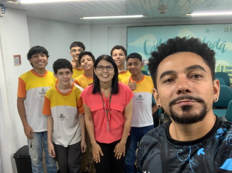 Selfie de um homem à frente de um grupo de estudantes adolescentes e uma mulher adulta. Os alunos vestem camisetas amarelas e brancas da rede municipal de Fortaleza, e todos estão sorrindo dentro de uma sala com cadeiras verdes ao fundo.