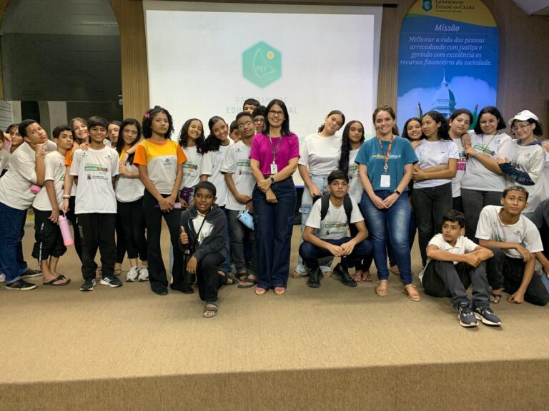 Grupo de cerca de 30 alunos do Projeto Integração da Univerdade Estadual do Ceará posando em um palco ou sala de conferências, em frente a uma tela de projeção com a logo do PEF e a Missão do Governo do Ceará em destaque. Os alunos, vestindo camisetas brancas, estão acompanhados por duas mulheres adultas ao centro, uma de blusa pink e outra de blusa azul-esverdeada.