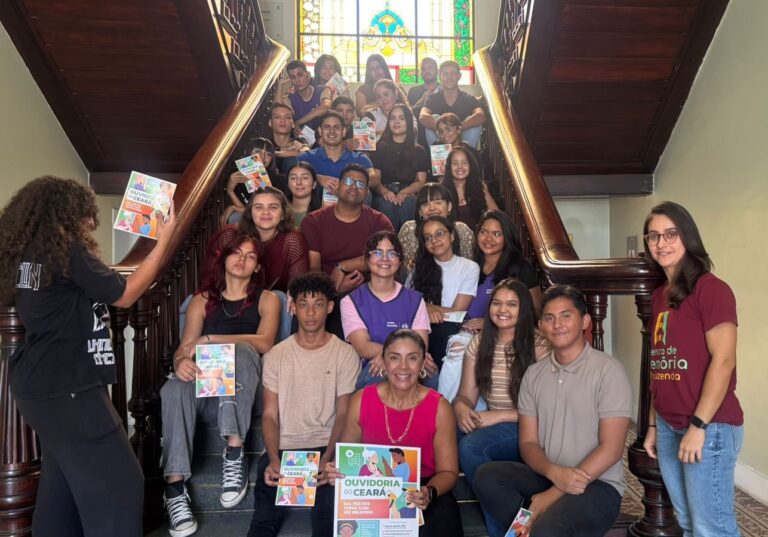 Grupo de jovens posa distribuído pela escadaria interna de um prédio histórico, com um grande vitral colorido ao fundo. A maioria segura folhetos coloridos da Ouvidoria do Ceará. Duas mulheres adultas estão posicionadas nas laterais da escadaria; uma delas usa camisa rosa e está sentada à frente do grupo.