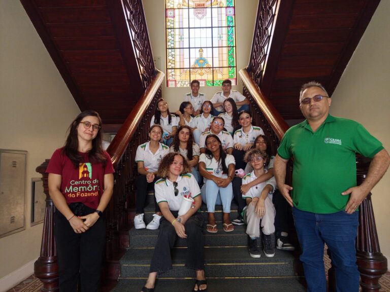 "Uma fotografia em grupo com 14 alunos e dois adultos na escadaria de madeira do Centro de Memória da Fazenda. Os estudantes vestem uniformes escolares brancos com detalhes verdes e o brasão do Governo do Ceará. À esquerda, uma mulher usa uma camiseta bordô com o logotipo do 'Centro de Memória da Fazenda'. À direita, o professor Francisco Freitas veste uma camisa polo verde. Ao fundo, no topo da escada, destaca-se um vitral colorido com a inscrição em latim 'Labor Omnia Vincit'."