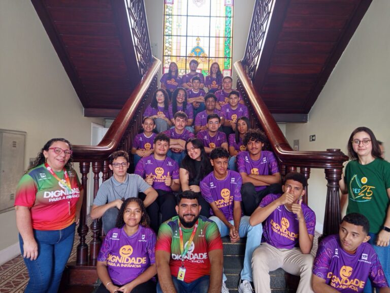 Grupo de estudantes e dois profissionais adultos posam sentados na escadaria interna de um prédio histórico, com vitral colorido ao fundo. Os alunos vestem camisetas roxas com a frase “Dignidade para a Infância”, e os adultos usam camisetas coloridas do mesmo projeto. Todos estão distribuídos pelos degraus, sorrindo ou olhando para a câmera em pose de registro.