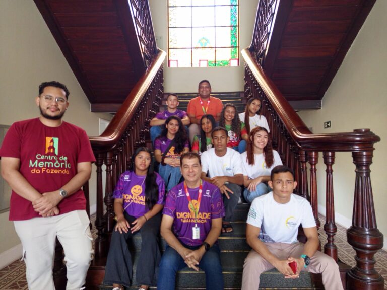 "Fotografia de um grupo de jovens da Associação Beneficente O Pequeno Nazareno acompanhados pelos professores José Ivan e Valdécio Batista. Eles estão posados em uma imponente escadaria de madeira clássica com um vitral colorido ao fundo. À esquerda, em destaque, um homem veste uma camiseta bordô com o logotipo 'Centro de Memória da Fazenda'. O grupo ocupa os degraus centrais, vestindo camisetas brancas, roxas e laranjas."