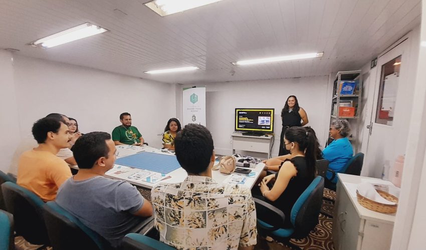 Participantes reunidos no Centro de Memória da Fazenda durante a oficina de restauração de fotografias, realizada pelo Museu Ferroviário. A museóloga Bruna Nunes está em pé à frente do grupo, explicando técnicas de preservação de fotografias. Algumas imagens e materiais de restauração estão sobre a mesa