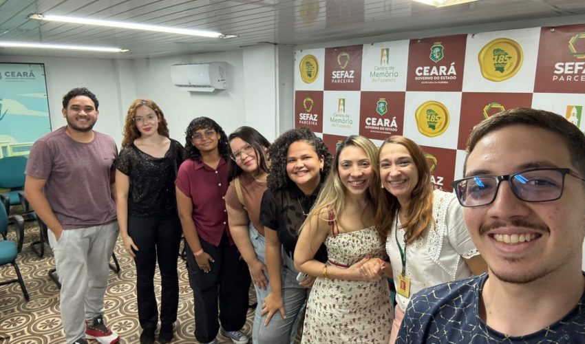 Na imagem, oito pessoas posam para uma selfie em uma sala, todas sorrindo em direção à câmera. O homem à direita segura o celular, enquanto os demais integrantes do grupo estão alinhados atrás dele. Ao fundo, vê-se um painel com as logomarcas do Governo do Estado do Ceará, da Sefaz e do Centro de Memória da Fazenda, além de um ar-condicionado fixado na parede. O chão possui um padrão geométrico em tons claros. Todos aparentam estar descontraídos e felizes, formando um ambiente acolhedor e amistoso