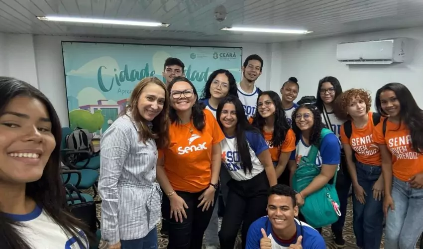 Grupo de 14 alunos do curso de Auxiliar Administrativo, acompanhados pela professora Cristiane, visitando o Centro de Memória da Fazenda. Os visitantes estão observando uma exposição ou atividade no espaço, com algumas cadeiras e elementos expositivos visíveis ao fundo