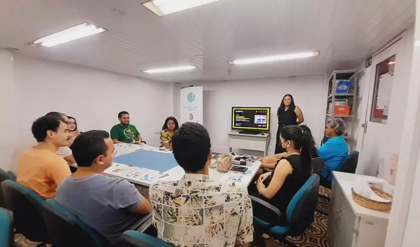 Participantes reunidos no Centro de Memória da Fazenda durante a oficina de restauração de fotografias, realizada pelo Museu Ferroviário. A museóloga Bruna Nunes está em pé à frente do grupo, explicando técnicas de preservação de fotografias. Algumas imagens e materiais de restauração estão sobre a mesa